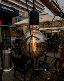 A close-up of a vintage-style light bulb hanging outdoors, surrounded by string lights. It is set against the backdrop of a hotel exterior with beige umbrellas and outdoor seating visible. The atmosphere suggests a cozy outdoor setting, possibly at a café or patio.