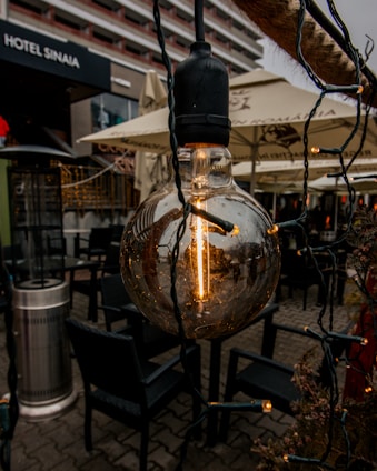 A close-up of a vintage-style light bulb hanging outdoors, surrounded by string lights. It is set against the backdrop of a hotel exterior with beige umbrellas and outdoor seating visible. The atmosphere suggests a cozy outdoor setting, possibly at a café or patio.