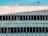 An abstract architectural facade featuring vertical and diagonal metal bars over the exterior of a building. The structure has a minimalist design with a pale color palette and a clear blue sky in the background.