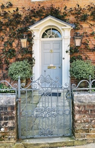 An ornate, wrought-iron gate stands in front of a classic brick building adorned with creeping ivy. A pair of symmetrical wall-mounted lanterns flank a light gray door that features the name 'Wynnflete House' above it. The door is framed by white columns and an arched transom window, creating an elegant entrance.