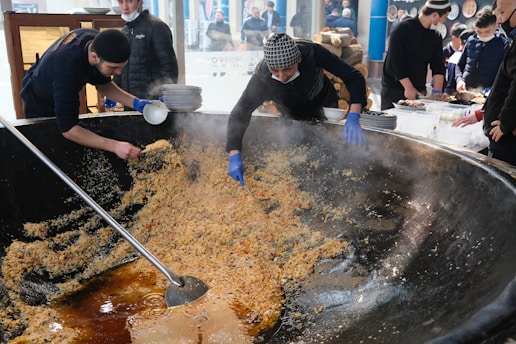 Several people are preparing a large dish in an enormous pan. Two people are actively stirring the food with large utensils, while others are assisting by holding plates and coordinating. The scene is bustling and suggests a communal cooking event.