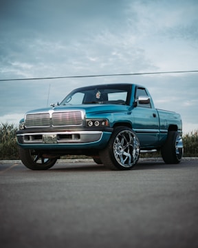 A modified turquoise pickup truck with large chrome rims is parked on a paved surface. The sky is overcast, creating a neutral background while tall grasses are visible in the distance. The truck features a customized front grille and a sleek, polished finish.