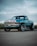 A modified turquoise pickup truck with large chrome rims is parked on a paved surface. The sky is overcast, creating a neutral background while tall grasses are visible in the distance. The truck features a customized front grille and a sleek, polished finish.