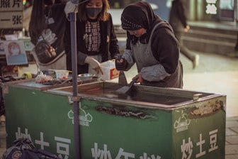 Two women are working at a street food stall. One of them is wearing a headscarf and apron, preparing food on a hotplate. The stall has a green front with white text and some displayed items. The scene is set in an urban area, possibly a market.