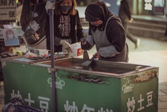 Two women are working at a street food stall. One of them is wearing a headscarf and apron, preparing food on a hotplate. The stall has a green front with white text and some displayed items. The scene is set in an urban area, possibly a market.