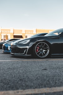 A black sports car is parked on a paved lot, next to a blue car. The sleek design includes glossy paint, stylish rims, and visible brake calipers. The background features a shopping center with store signs visible.