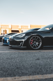A black sports car is parked on a paved lot, next to a blue car. The sleek design includes glossy paint, stylish rims, and visible brake calipers. The background features a shopping center with store signs visible.