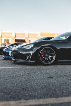 A black sports car is parked on a paved lot, next to a blue car. The sleek design includes glossy paint, stylish rims, and visible brake calipers. The background features a shopping center with store signs visible.