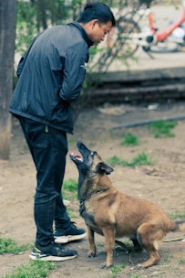 A man wearing a black jacket and dark jeans stands outdoors facing a brown dog. The dog is sitting attentively and looking up at the man. The background is a park-like area with a dirt path and patches of grass.