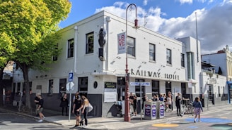 A white two-story building labeled 'Railway Hotel' features a classic architectural design with several people gathered outside. A bright red lamppost with a 'Love local' sign stands in front, alongside a leafy tree casting shade over the sidewalk. The street is lively with pedestrians, and there are signs and posters on the walls. The sky is partly cloudy, adding a dynamic background.
