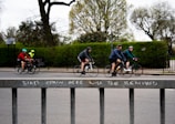 A group of cyclists ride along a street, each wearing helmets and colorful athletic clothing. In the foreground, a metal fence displays a warning message: 'BIKES CHAIN HERE WILL BE REMOVED.' Leafless trees and green shrubs are visible in the background, suggesting a park-like setting.