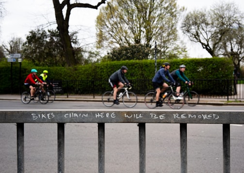 A group of cyclists ride along a street, each wearing helmets and colorful athletic clothing. In the foreground, a metal fence displays a warning message: 'BIKES CHAIN HERE WILL BE REMOVED.' Leafless trees and green shrubs are visible in the background, suggesting a park-like setting.
