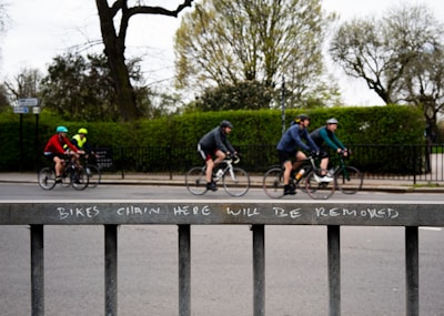 A group of cyclists ride along a street, each wearing helmets and colorful athletic clothing. In the foreground, a metal fence displays a warning message: 'BIKES CHAIN HERE WILL BE REMOVED.' Leafless trees and green shrubs are visible in the background, suggesting a park-like setting.