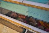 Two individuals, possibly a mother and child, are asleep on a concrete surface. They are seen through a transparent barrier with text and pipes running horizontally across it. The woman wears a colorful sari, and her child is nestled closely to her. The environment appears to be a street or public area.