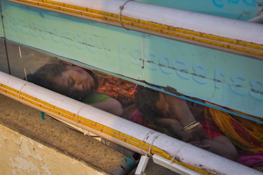 Two individuals, possibly a mother and child, are asleep on a concrete surface. They are seen through a transparent barrier with text and pipes running horizontally across it. The woman wears a colorful sari, and her child is nestled closely to her. The environment appears to be a street or public area.