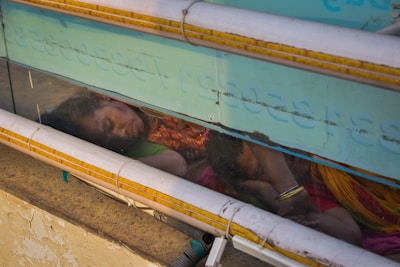 Two individuals, possibly a mother and child, are asleep on a concrete surface. They are seen through a transparent barrier with text and pipes running horizontally across it. The woman wears a colorful sari, and her child is nestled closely to her. The environment appears to be a street or public area.