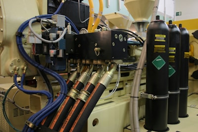 Complex industrial machinery with multiple interconnected hoses and pipes, featuring control panels and metallic cylinders. The setup includes blue and yellow hoses, with metallic fastenings and electrical components.