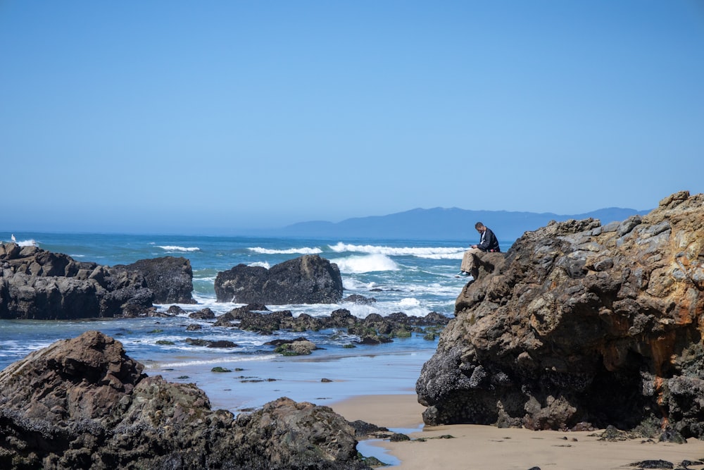 person in black shirt sitting on rock formation near sea during daytime