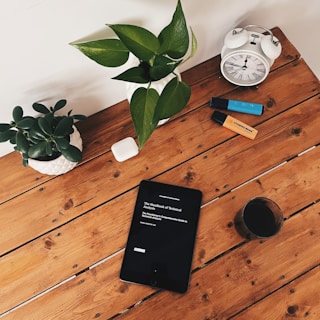 A wooden table holds an assortment of objects including two potted plants, an alarm clock, a pair of markers, a tablet displaying a book, a small white electronic device, and a glass of dark liquid. The tablet's screen shows the title 'The Handbook of Technical Analysis'.