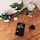 A wooden table holds an assortment of objects including two potted plants, an alarm clock, a pair of markers, a tablet displaying a book, a small white electronic device, and a glass of dark liquid. The tablet's screen shows the title 'The Handbook of Technical Analysis'.