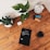 A wooden table holds an assortment of objects including two potted plants, an alarm clock, a pair of markers, a tablet displaying a book, a small white electronic device, and a glass of dark liquid. The tablet's screen shows the title 'The Handbook of Technical Analysis'.