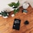A wooden table holds an assortment of objects including two potted plants, an alarm clock, a pair of markers, a tablet displaying a book, a small white electronic device, and a glass of dark liquid. The tablet's screen shows the title 'The Handbook of Technical Analysis'.
