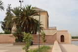A traditional building with Moroccan architectural elements stands beside a pathway, partially obscured by lush palm trees and other green vegetation. The structure has a flat roof and decorative tile work, with a simple wooden door set into a peach-colored wall. The scene is tranquil and seems to be set in an outdoor public or historical area.