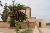 A traditional building with Moroccan architectural elements stands beside a pathway, partially obscured by lush palm trees and other green vegetation. The structure has a flat roof and decorative tile work, with a simple wooden door set into a peach-colored wall. The scene is tranquil and seems to be set in an outdoor public or historical area.
