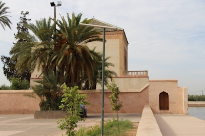 A traditional building with Moroccan architectural elements stands beside a pathway, partially obscured by lush palm trees and other green vegetation. The structure has a flat roof and decorative tile work, with a simple wooden door set into a peach-colored wall. The scene is tranquil and seems to be set in an outdoor public or historical area.
