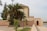 A traditional building with Moroccan architectural elements stands beside a pathway, partially obscured by lush palm trees and other green vegetation. The structure has a flat roof and decorative tile work, with a simple wooden door set into a peach-colored wall. The scene is tranquil and seems to be set in an outdoor public or historical area.