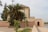 A traditional building with Moroccan architectural elements stands beside a pathway, partially obscured by lush palm trees and other green vegetation. The structure has a flat roof and decorative tile work, with a simple wooden door set into a peach-colored wall. The scene is tranquil and seems to be set in an outdoor public or historical area.