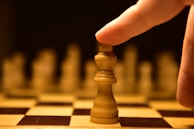 A hand poised to move a king chess piece on a chessboard, with other pieces blurred in the background. The lighting creates a dramatic and focused atmosphere on the central piece.