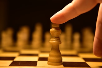 A hand poised to move a king chess piece on a chessboard, with other pieces blurred in the background. The lighting creates a dramatic and focused atmosphere on the central piece.