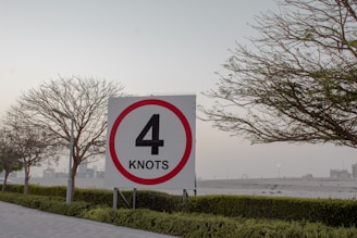 A speed limit sign displaying the number 4 in bold black text against a white background with a red circular border. It is placed in an outdoor setting, surrounded by trees with sparse foliage, neatly trimmed bushes, and a paved walkway. The sky appears overcast, with urban buildings visible in the distance.