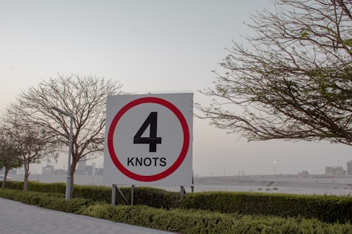 A speed limit sign displaying the number 4 in bold black text against a white background with a red circular border. It is placed in an outdoor setting, surrounded by trees with sparse foliage, neatly trimmed bushes, and a paved walkway. The sky appears overcast, with urban buildings visible in the distance.