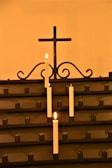 A row of white candles is lit, placed in holders on a dark metal stand. The background features a simple metal cross with decorative scrollwork beneath it. The overall ambiance is warm and serene, highlighted by the soft glow of the candlelight.