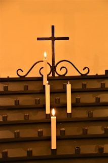 A row of white candles is lit, placed in holders on a dark metal stand. The background features a simple metal cross with decorative scrollwork beneath it. The overall ambiance is warm and serene, highlighted by the soft glow of the candlelight.