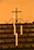 A row of white candles is lit, placed in holders on a dark metal stand. The background features a simple metal cross with decorative scrollwork beneath it. The overall ambiance is warm and serene, highlighted by the soft glow of the candlelight.