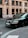 A black luxury car is parked on a city street. The car's brand logo is visible, and it's positioned near a building with large windows. There's a street sign and a bicycle in the background.