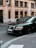 A black luxury car is parked on a city street. The car's brand logo is visible, and it's positioned near a building with large windows. There's a street sign and a bicycle in the background.