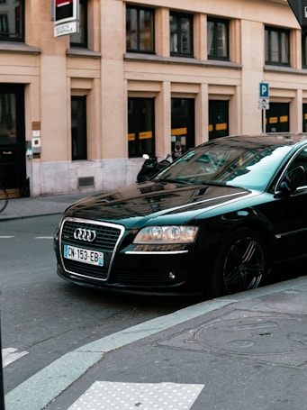 A black luxury car is parked on a city street. The car's brand logo is visible, and it's positioned near a building with large windows. There's a street sign and a bicycle in the background.