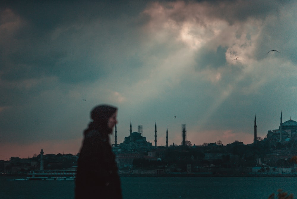 woman in black dress standing near body of water under cloudy sky during daytime