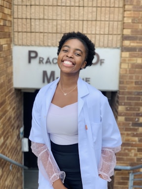 A person wearing a white lab coat and a white top stands smiling in front of a brick wall. The wall has signage partially visible, possibly related to a medical practice. The person exudes confidence and professionalism.