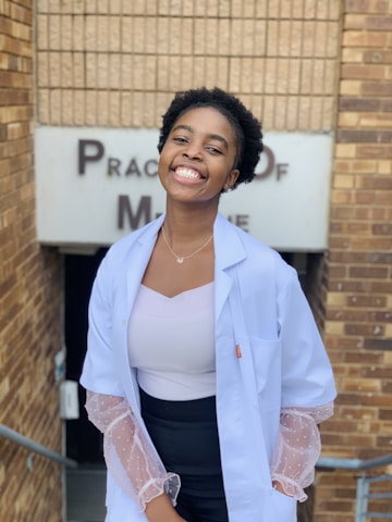 A person wearing a white lab coat and a white top stands smiling in front of a brick wall. The wall has signage partially visible, possibly related to a medical practice. The person exudes confidence and professionalism.