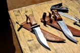Two large hunting knives with wooden handles and shiny steel blades resting in brown leather sheaths are laid on a wooden table. Near the knives, there is a square tool and a metal ruler. The table appears to be outdoors, indicated by natural lighting and shadows.