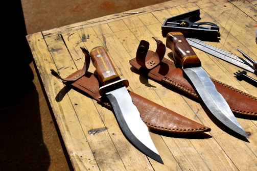 Two large hunting knives with wooden handles and shiny steel blades resting in brown leather sheaths are laid on a wooden table. Near the knives, there is a square tool and a metal ruler. The table appears to be outdoors, indicated by natural lighting and shadows.