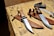 Two large hunting knives with wooden handles and shiny steel blades resting in brown leather sheaths are laid on a wooden table. Near the knives, there is a square tool and a metal ruler. The table appears to be outdoors, indicated by natural lighting and shadows.