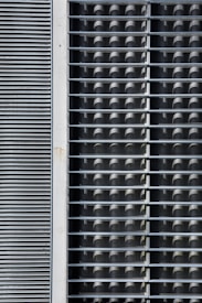 The image features a geometric pattern created by overlapping rectangular and circular shapes. The left side consists of horizontal slats arranged closely, resembling a vent or louver. The right side shows a grid of circular tubes or pipes stacked within a metal framework.