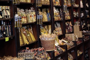 A variety of packaged Italian foods, including pasta and olive oil, are arranged neatly on dark wooden shelves in a store. There are several types of pasta in clear bags, bottles of olive oil with different labels, and a basket filled with wooden utensils. Some shelves also contain jars of sauces and other food items, alongside wine bottles.