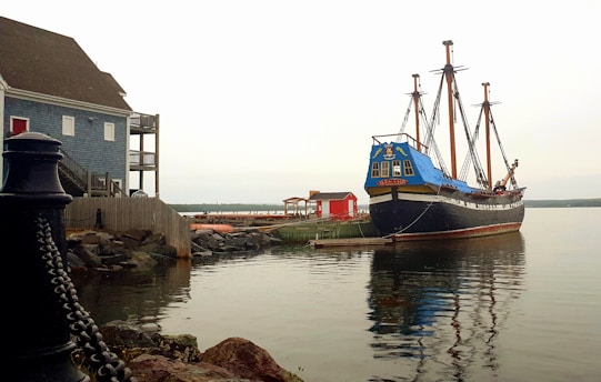 A large, wooden ship is docked near the shore, with its tall masts and rigging visible. The ship, painted in shades of blue and black, is settled on calm waters. Nearby, a blue building with a red door and a wooden deck is situated on the rocky shore. In the background, there is a small red structure and a distant treeline under an overcast sky.